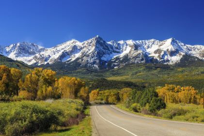 Picturesque landscape, wide open road, snowcapped mountains in distance, blue skies overhead. 