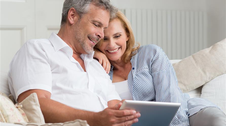 A mature couple cuddling on couch while looking at a tablet. 
