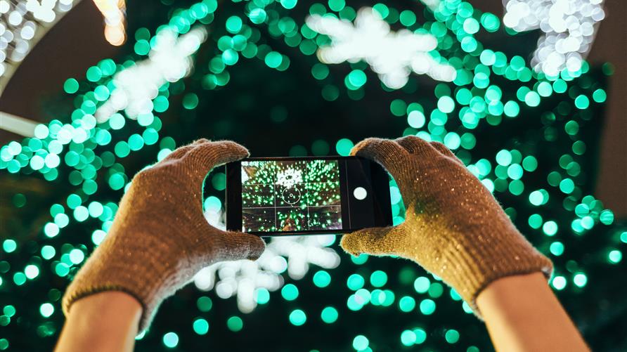 Close up shot of a woman in gloves taking pictures of a Christmas light display with a smart phone. 