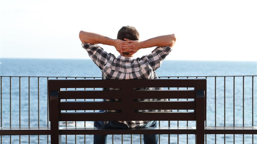 Man relaxing with his hands behind his neck on a bench overlooking the ocean. 
