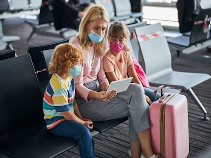 Mother will two small children wearing face coverings looking at a tablet at the airport. 