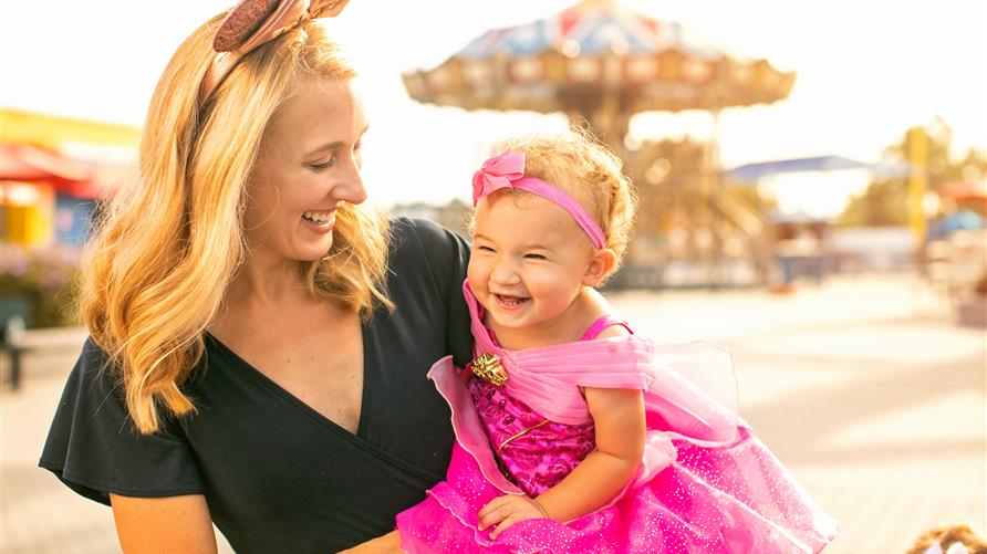 Mother wearing sparkly Minnie Mouse ears smiling and holding young daughter wearing a Disney Princess costume. 