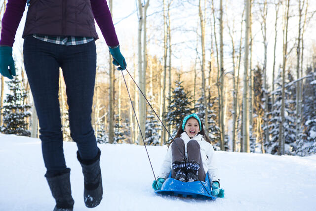 Adult woman pulling young girl on a sled through the snow while on vacation at Sunrise Lodge, a Hilton Grand Vacations Club in Park City, Utah. 
