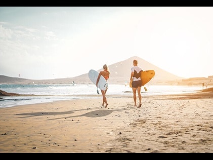 Couple walking together out to sea with surf boards in Maui with mountains in the distance. 