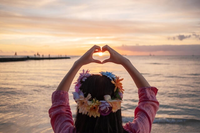 Woman wearing a flower crown and making heart hands toward the sunset in Hawaii. 