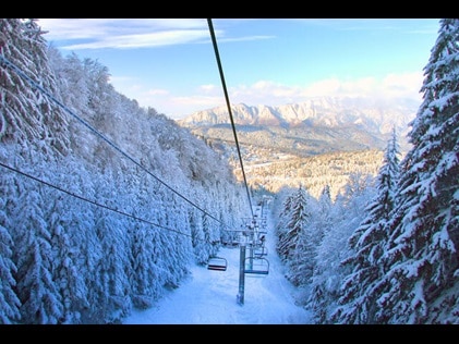  Snow covered ski lift with evergreen trees on either side and picturesque mountains in the distance ahead. 