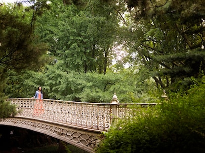 Woman walking across Central Park bridge surrounded by lush greenery, New York, New York. 