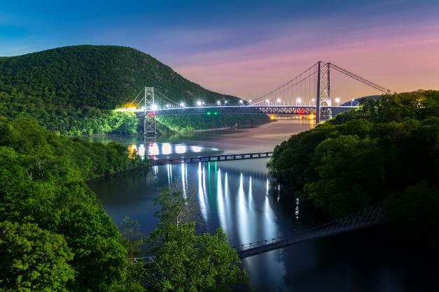 Aerial shot of Bear Mountain Bridge illuminated at dusk with purple sunset skies overhead, New York. 