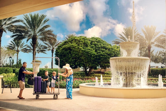 Family of four checking-in at with a luggage cart in front of fountain at Tuscany Village, a Hilton Grand Vacations Club, Orlando. 