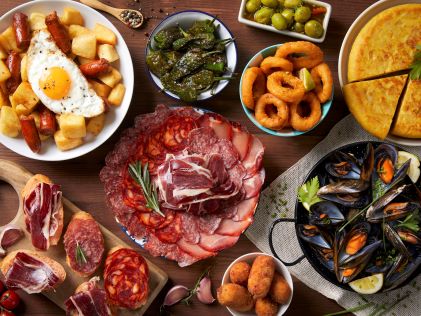 An overhead view of a wooden table with several Spanish tapas dishes, including ham, mussels and peppers