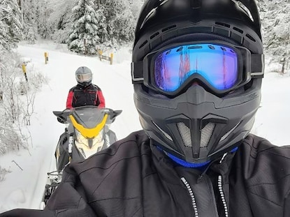 Hilton Grand Vacations Owners snapping selfie, snowmobiling, snow-covered trees trail, trees in distance. 