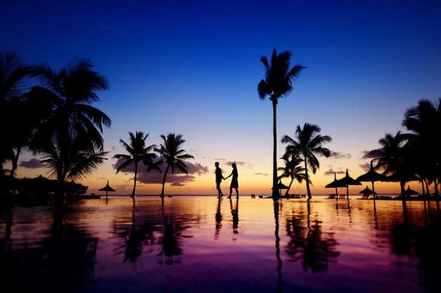 Silouhette of couple walking hand in hand along the edge of palm tree-lined infinity pool at sunset, Maldives.
