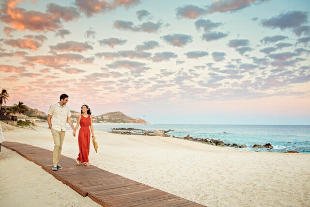 Couple strolling hand in hand along oceanfront wooden walkway, sunset painted skies overhead, Los Cabos, Mexico.