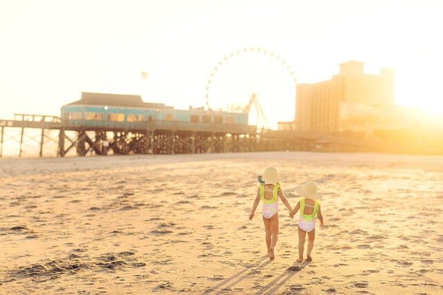 Two young girls walking hand in hand along beach at sunset, SkyWheel and boardwalk in distance, Myrtle Beach, South Carolina.