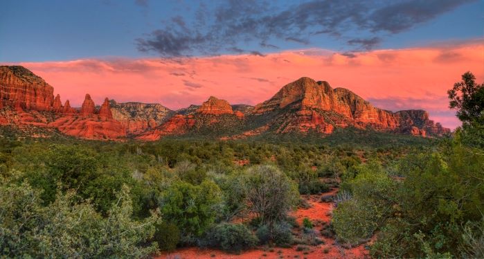 The desert near Sedona, Arizona. There are red rock formations in the mid-ground, a blue and orange sunset in the background and green desert shrubs in the foreground. 