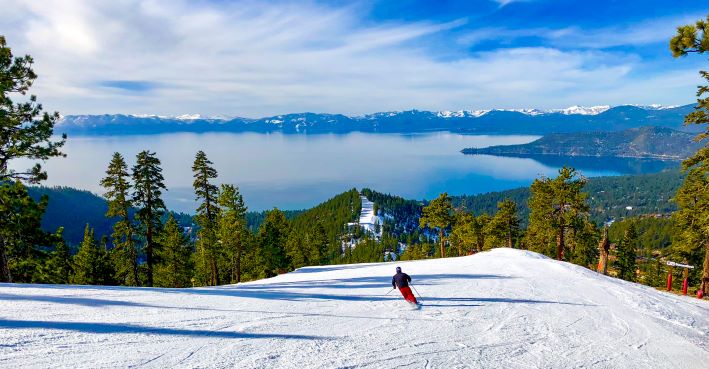Traveler skiing downhill towards Lake Tahoe, tree-lined ski slope, blue skies overhead and mountains in the distance, Nevada. 