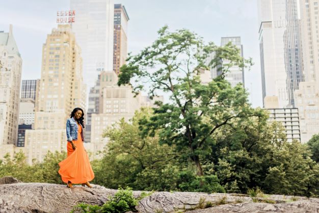 A woman in an orange dress poses in Central Park, New York, skyscrapers visible behind her