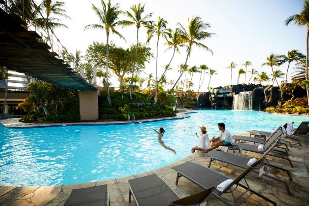 A family relaxes by the pool at the Hawaiian Waikoloa Village resort as a child jumps in, palm trees in the background