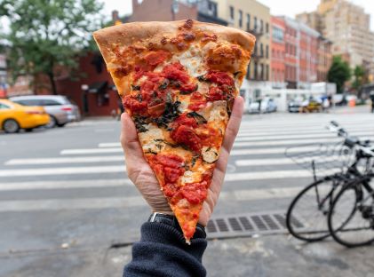 A hand holds up a slice of pizza against a background of a New York City street