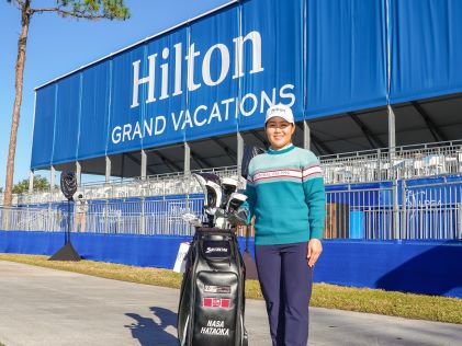 Nasa Hataoka, a Hilton Grand Vacations celebrity Brand Ambassador, standing with her golf bag in front of a sign for the Hilton Grand Vacations Tournament of Champions