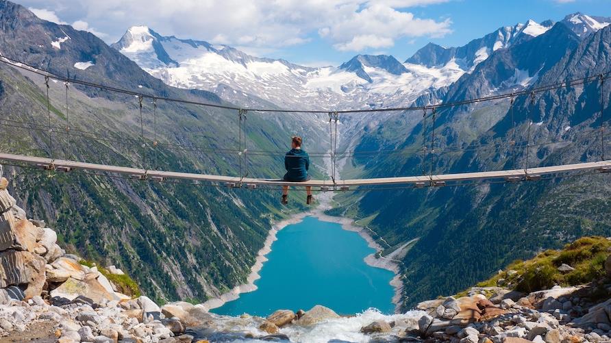 Gorgeous aerial shot of a man sitting on a suspended bridge over a river canyon with snow capped mountains in the distance