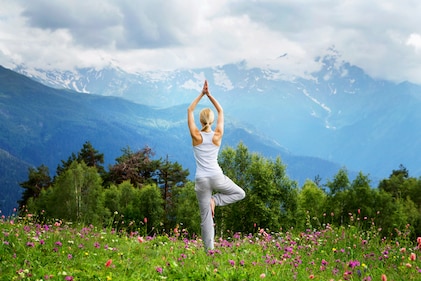 A woman stands in yoga tree pose overlooking a field of flowers with mountains on the horizon