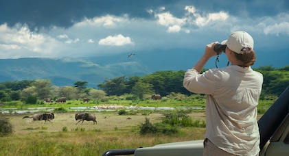A female tourist on safari in Africa using binoculars to watch wildebeest and elephants in the wild savanna, Manyara National Park
