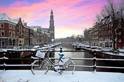 Beautiful winter scene, snow-covered bicycle, historic buildings lining urban canal, cotton-candy skies overhead, Amsterdam, Netherlands.
