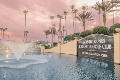 Mystic Dunes, a Hilton Vacation Club resort in Orlando. Photo features the entrance and fountain in the foreground, palm trees in the distance and pink clouds above.