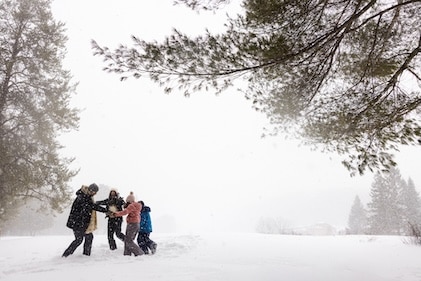 Family playing and holding hands in a circle in a field of snow under snowy skies and some pine trees at Tremblant, a Hilton Grand Vacation Club in Quebec, Canada.