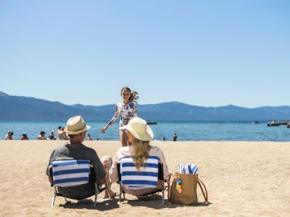 Two adults relax in striped lounge chairs on the beach while a child runs to play in the water