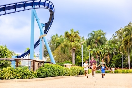 Family of four enjoys snacks as they walk through a theme park with large rollercoaster to the left of the frame.