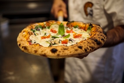 Close up of a chef holding a fire-roasted pizza and lots of toppings.