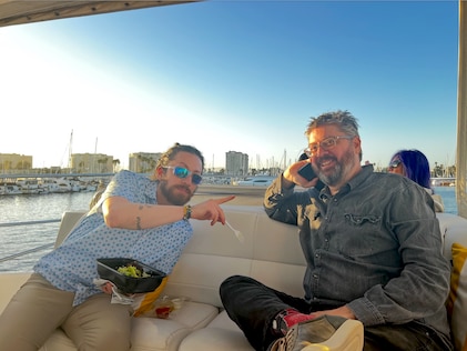 Two men enjoying time out on a speedboat on a sunny day with one eating a salad and the other on his cellphone. Both are smiling and one is leaning in toward the other for the photo.