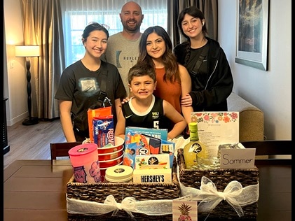 Young family of 5 poses facing the camera inside Marbrisa, a Hilton Grand Vacations Club with two very large gift baskets displaying treats, wine and gift cards.