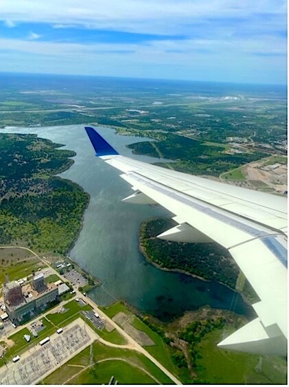 View out of the window of a plane flying over Austin Texas, showing wingspan with the land and a river far below.