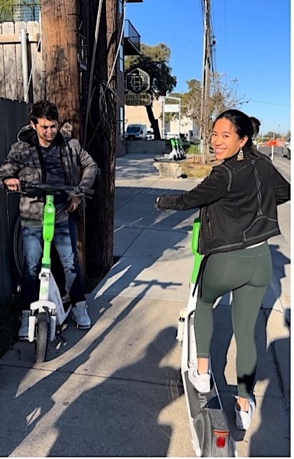 View of young man and woman looking over her shoulder on bright green scooters as they travel down a sidewalk in Austin.