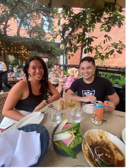 A smiling young couple dining on a restaurant patio with plates of food in front of them and trees behind them against a red brick building.