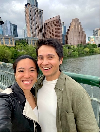 Young couple smiling at the camera as they pose on the Congress Avenue Bridge with the Austin Texas in the far background.