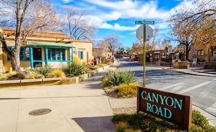 Image of the sunny street along Canyon Road in Santa Fe, New Mexico showing art galleries and tree-lined sidewalks and the Canyon Road sign in the foreground.