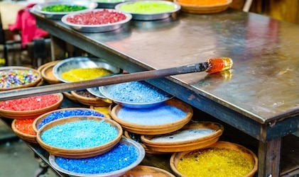 Colorful plates of blue, yellow red and orange glass bits in a glassmaking studio as well as a worktable and pole with heated glas at the tip as it's being shaped.