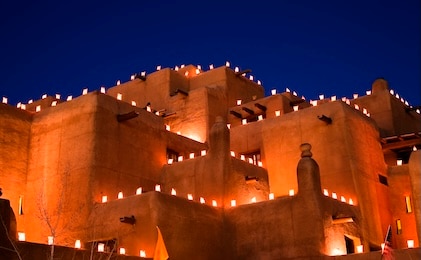 A series of adobe-style rooftops lit by candles under a night sky in Santa Fe, New Mexico.