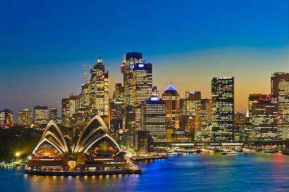 Tall buildings and the Sydney Opera house seen from the harbor at dusk. The lights in the skyscrapers and opera house cast a golden glow, and the remnants of the sunset are on the horizon.