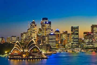 Tall buildings and the Sydney Opera house seen from the harbor at dusk. The lights in the skyscrapers and opera house cast a golden glow, and the remnants of the sunset are on the horizon.