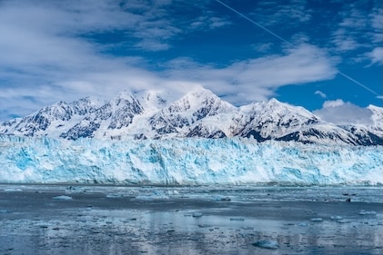 View of Hubbard Glacier in Alaska from across the water. In the background are snow-capped mountain range against blue skies.