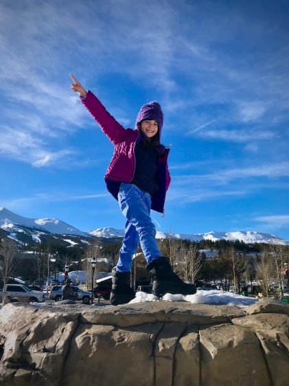 A child balances on a snowy rock in winter gear while pointing up at the sky