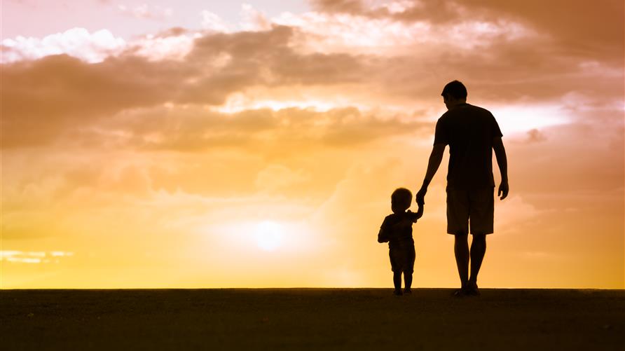 Silhouette of a father and young child holding hands and walking into the sunset. 