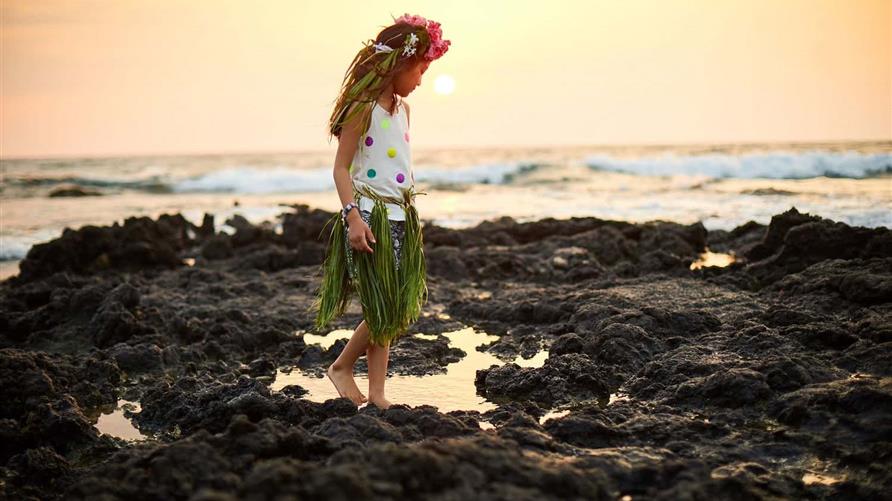 Little girl in grass skirt and lei exploring the rocks along the Pacific shoreline in Hawaii. 