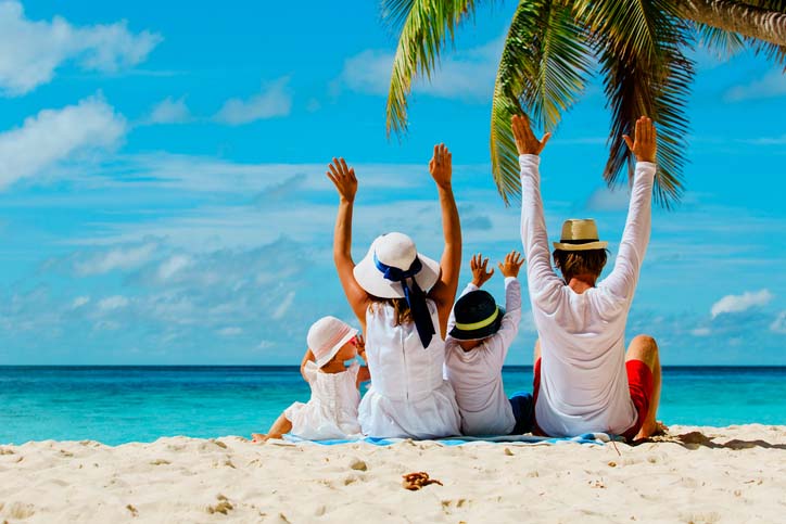 A family sitting under a palm tree looking out at the ocean with arms raised in excitement. 