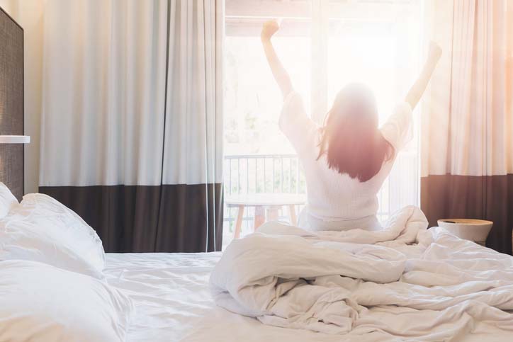 Woman stretching in bed in a Hilton Hotel room looking at the sunrise outside the window.